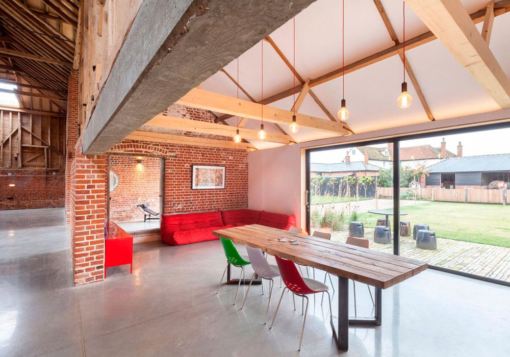 Modern open-plan living space featuring large sliding glass doors, wooden beams, red seating, and dining table with garden views beyond.