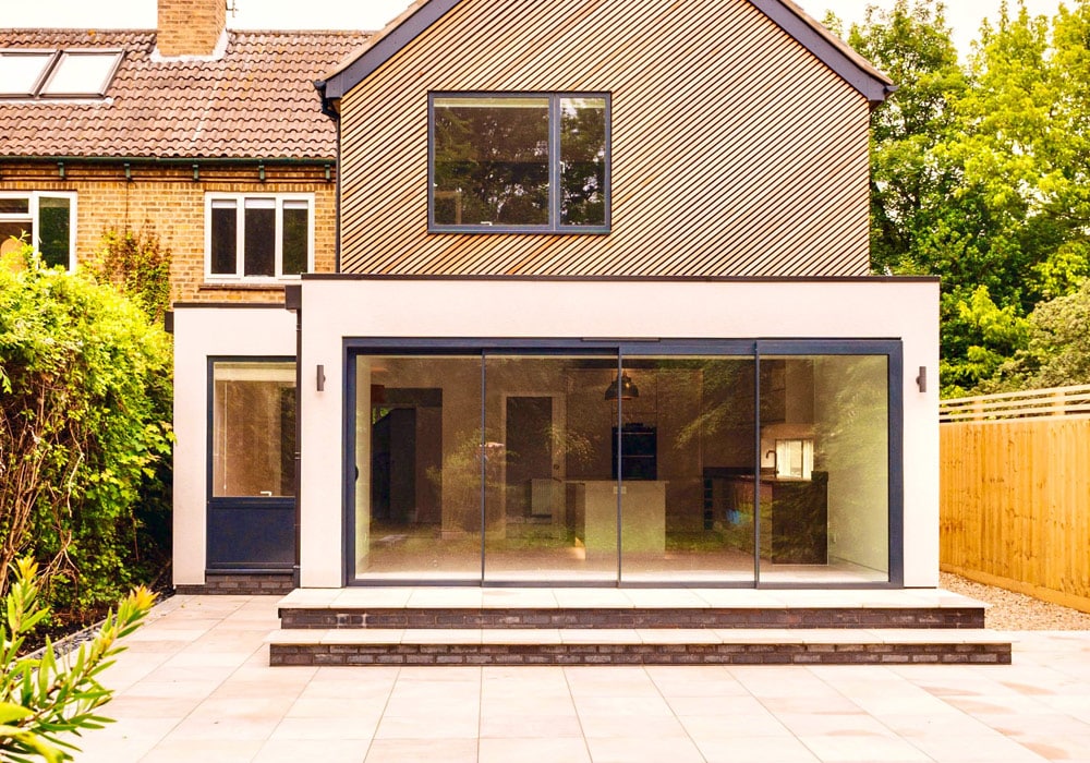 Modern white extension with large sliding glass doors opening onto a patio, flanked by lush greenery.