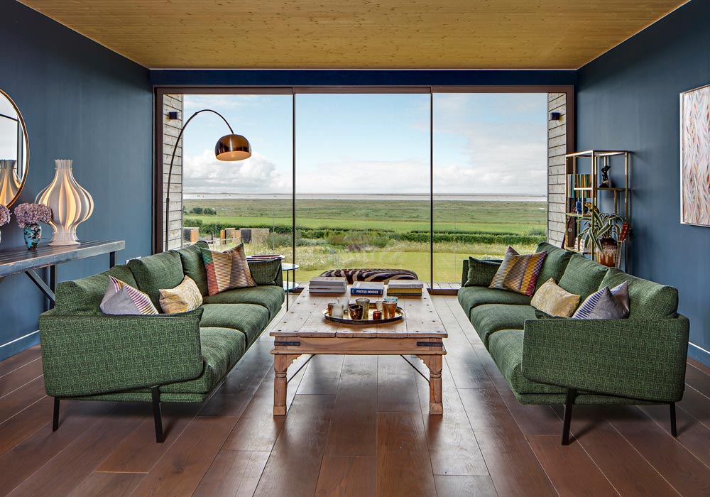Modern living room with aluminium sliding doors opening onto expansive countryside views, featuring green curved armchairs and wooden flooring.