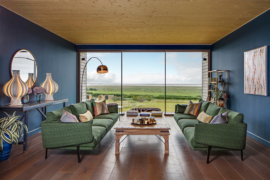 Modern living room with expansive glazed doors framing countryside views, featuring green armchairs and wooden flooring, exemplifying IDSystems premium glazing solutions.