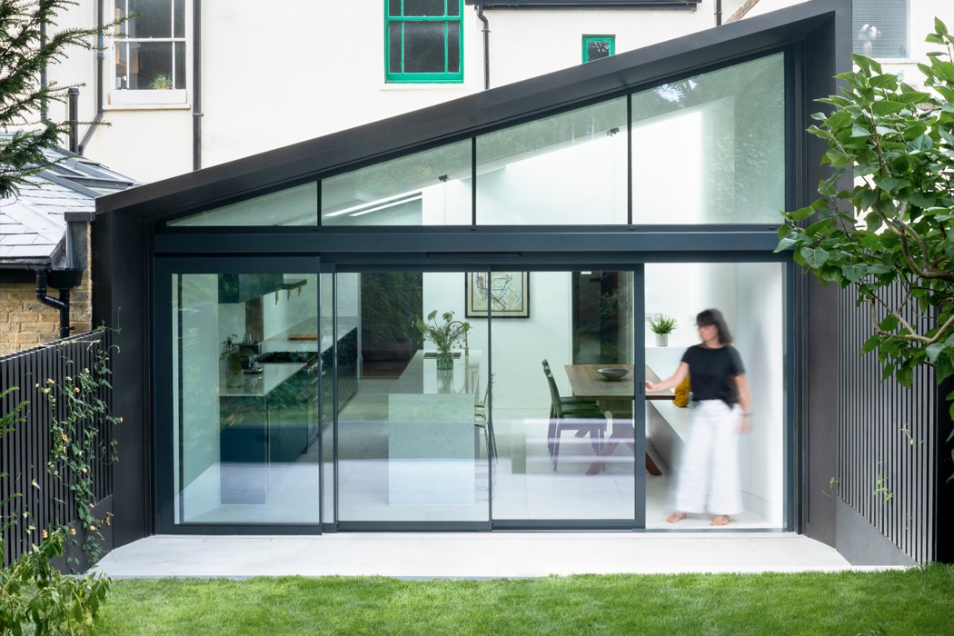 Modern black-framed double ultra-thin theEdge2 sliding doors open onto a garden, revealing a bright contemporary kitchen-dining space with a woman inside.