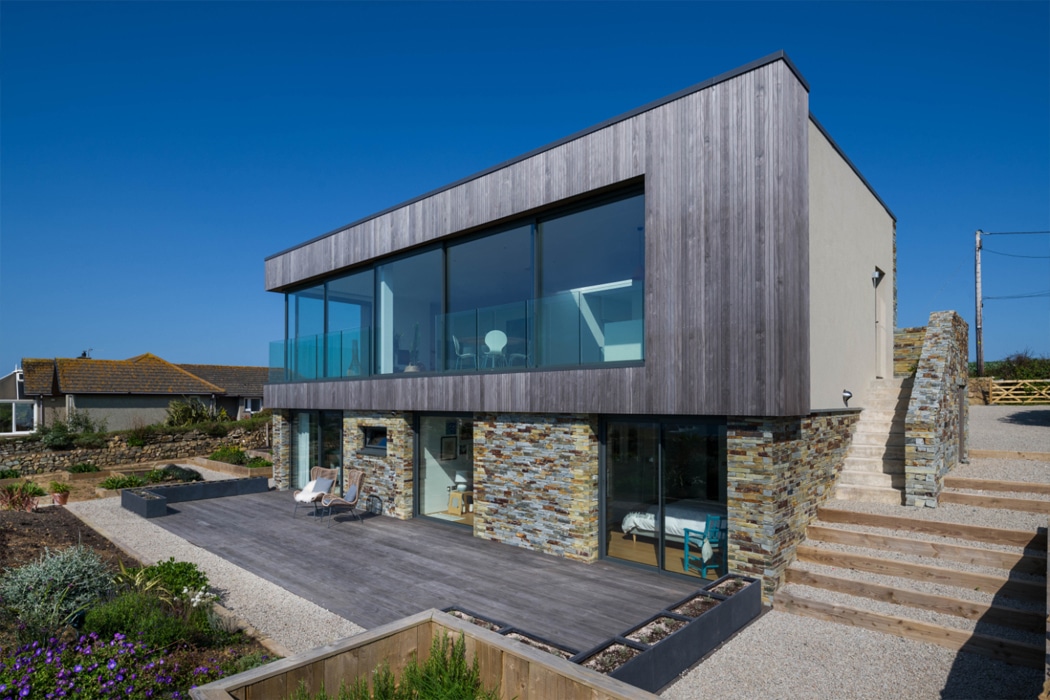 Modern stone house with theEDGE2 ultra-slimline glass sliding doors opening onto a dark composite deck, showcasing expansive glazing and seamless indoor-outdoor living.
