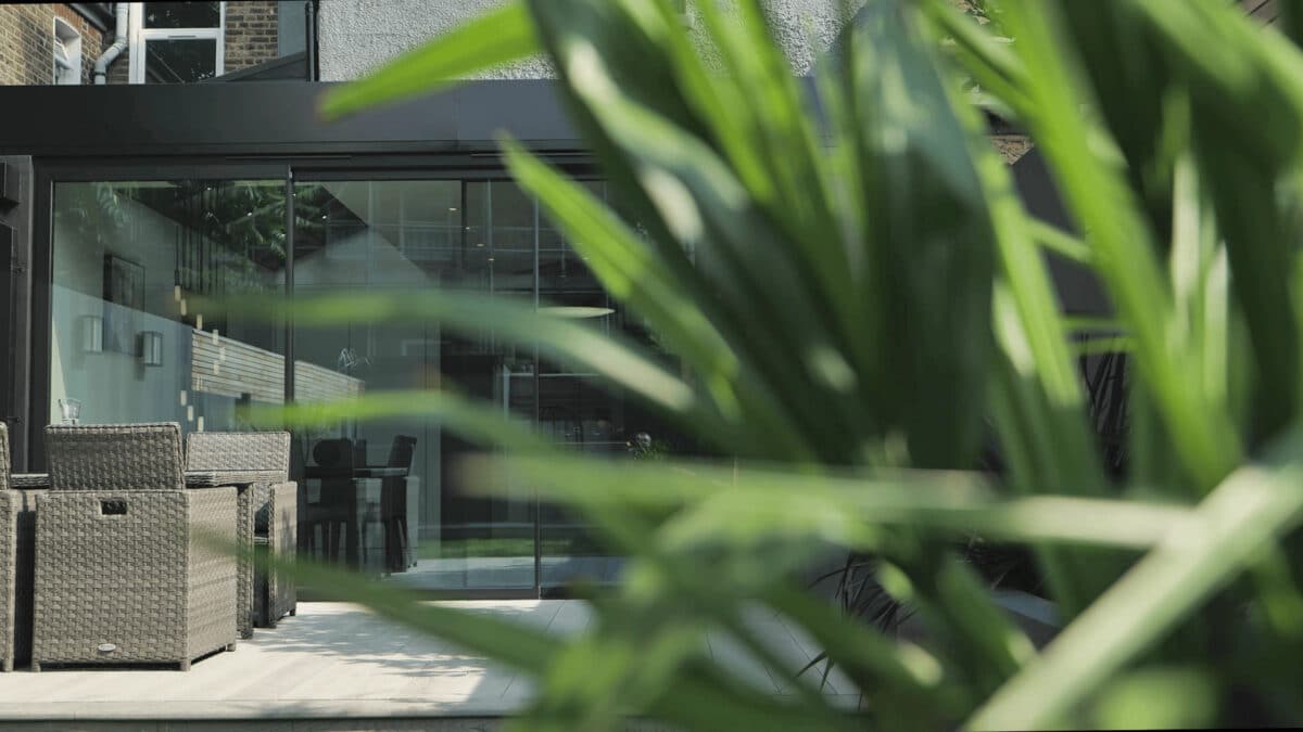 Modern grey house extension with slim-profile aluminium sliding doors opening onto a sunny patio, featuring contemporary outdoor furniture and ornamental grasses.