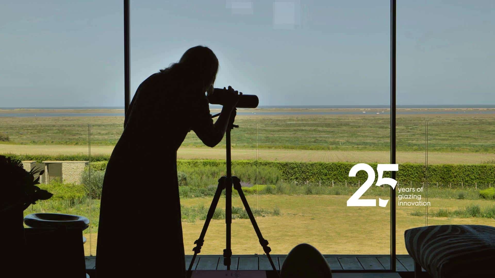 Silhouetted figure viewing expansive countryside through large glazed bifold doors and windows, showcasing panoramic views and natural light.